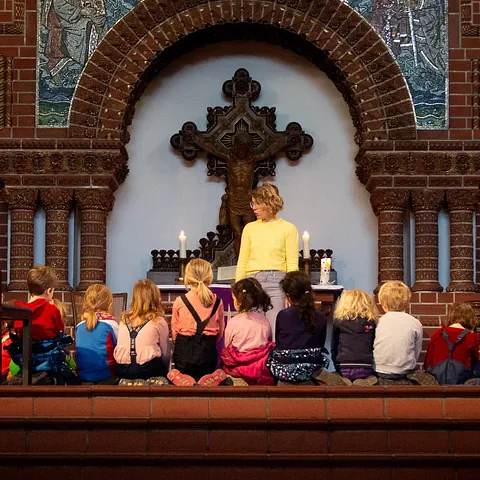 Kinder sitzen vor einer Frau und einem Kruzifix in einer Kirche.