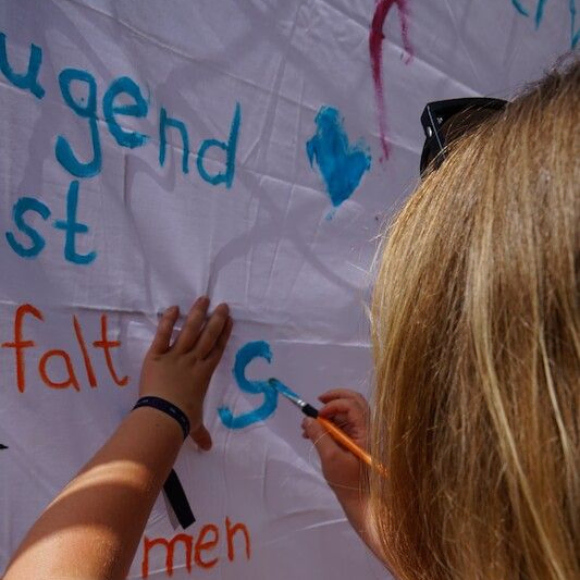 Eine blonde Person malt blaue Buchstaben auf ein weißes Stoffbanner